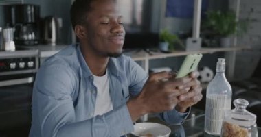 Akıllı telefon kullanan Afro-Amerikalı adam evde kahvaltı yaparken online içerikten keyif alıyor. Modern cihaz ve yaşam tarzı konsepti.