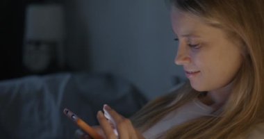 Neşeli genç bir kadın akıllı telefon ekranına dokunuyor gecenin karanlığında yatakta oturmuş gülümsüyor. İnternet iletişimi ve cihaz bağımlılığı kavramı.