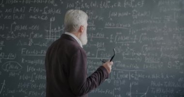Düşünceli kıdemli profesör formüllü karatahtaya bakıyor elinde gözlüklerle üniversitede bilimi düşünüyor. Eğitim ve bilgi kavramı.