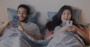 Kız ve erkek mutlu çift akıllı telefonlar kullanıyor ve sohbet ediyorlar akşamları yatakta uzanıyorlar. İlişki ve aygıt kavramı.