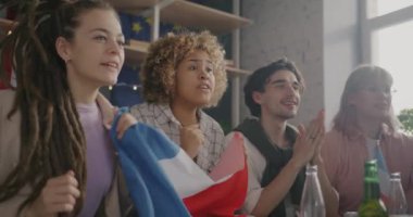TV 'de spor müsabakaları izleyen ve evde eğlenen Fransız bayrakları sallayan aşırı neşeli genç hayranların ağır çekimleri. Arkadaşlar ve eğlence konsepti.