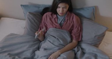 Uzaktan kumandayla televizyon seyreden neşeli genç bir kadın akşamları yatakta eğleniyor. Televizyon ve gençlik kültürü kavramı.