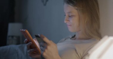 Akıllı telefon kullanan ve elinde kredi kartı tutan mutlu genç bir kadın akşamları yatakta alışveriş yapıyor. E-ticaret ve para kavramı.