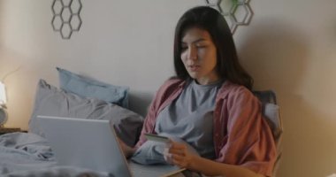 Diz üstü bilgisayarı ve kredi kartıyla alışveriş yapan neşeli Asyalı bayan akşam yatakta mutlu bir şekilde gülümsüyor. Modern teknoloji ve finans kavramı.