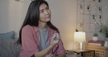 Genç Asyalı bayan televizyonda uzaktan kumandayla eğlenceli bir program izliyor. Yatak odasında tek başına eğleniyor. Kitle iletişim araçları ve boş zaman etkinliği kavramı.