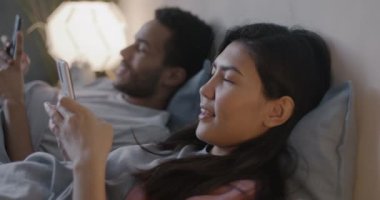 Mutlu çift Asyalı kadın ve Afrikalı Amerikalı erkek akıllı telefon kullanıyor ve akşamları yatakta uzanıp konuşuyorlar. İlişki ve modern teknoloji kavramı.