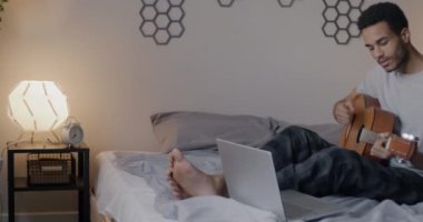Genç Afro-Amerikalı adam gitar çalmayı öğreniyor. Dizüstü bilgisayarla ders izlemeyi öğreniyor. Akşamları yatakta oturuyor. Müzik aleti ve gençlik hobisi.