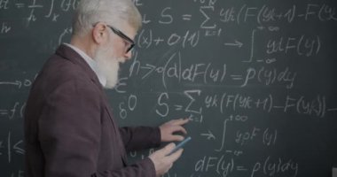 Üst düzey bilim adamı karatahtadaki matematik formülünü kontrol ediyor ve akıllı telefon uygulaması kullanıyor. Modern teknoloji ve bilim gelişimi kavramı.