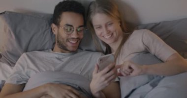 Kafkasyalı mutlu bir çift ve Afro-Amerikalı bir adam birlikte akıllı telefon kullanıyor ve akşamları yatakta uzanıp konuşuyorlar. Romantik ilişki ve alet kavramı.