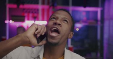 Neşeli Afrikalı Amerikalı adam dans ediyor ve cep telefonuyla konuşuyor gürültülü bir odada, gece müzik ve kalabalıkla. İletişim ve parti konsepti.