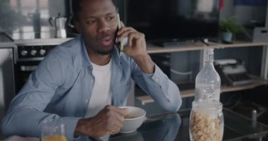 Neşeli Afrikalı Amerikalı adam kahvaltı yapıyor ve cep telefonuyla konuşuyor. Mutfaktaki masada oturuyor. İletişim ve beslenme kavramı.