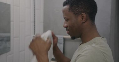 Afro-Amerikalı bir adam sabah banyoda yıkandıktan sonra ıslak yüzünü temiz beyaz havluyla siliyor. Günlük rutin ve hijyen konsepti.