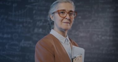 Üniversite sınıfında elinde kitaplarla kameraya bakan yaşlı kadın matematikçinin yavaş çekim portresi. Yüksek öğrenim ve bilim konsepti.