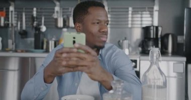 Gamsız Afrikalı Amerikalı bir adam akıllı telefonuyla mesajlaşıyor kahvaltıda evde oturuyor. Modern teknoloji ve evdeki insanlar.