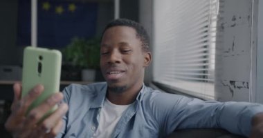 Neşeli Afro-Amerikalı adam akıllı telefonuyla çevrimiçi video konuşması yapıyor dairedeki koltukta otururken görüntüye bakıyor. İletişim ve insan kavramı.