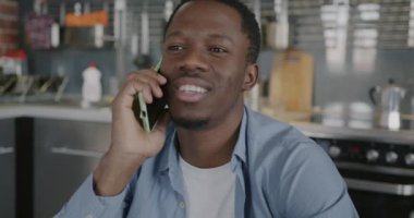 Neşeli Afro-Amerikalı adam cep telefonuyla sohbet ediyor. Mutfakta oturuyor. Modern teknoloji ve iletişim konsepti.