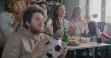 TV 'de futbol maçı izleyen heyecanlı taraftarların yavaş çekimi ve evde eğlenmeyi destekleyen Amerikan bayrağı. Spor ve gençlik kavramı.