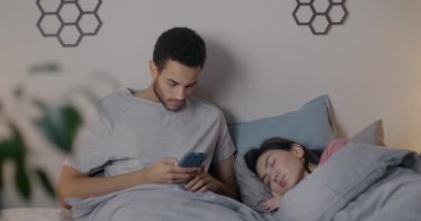 Akşamları karısı yatakta uyurken akıllı telefon kullanan Afrikalı Amerikalı bir adam sosyal medyanın tadını çıkarıyor. Aile yaşam tarzı ve modern teknoloji kavramı.