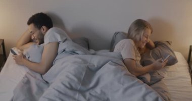 Karı koca akıllı telefonları kullanıyor. Birbirlerini görmezden geliyorlar. Yatağın farklı taraflarında yatıyorlar. Evlilik ve modern teknoloji kavramı.