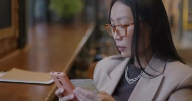 Akıllı telefon ve kredi kartı alışverişi yapan Asyalı iş kadını kafede elektronik parayla ödeme yapıyor. Modern teknoloji ve finans kavramı.