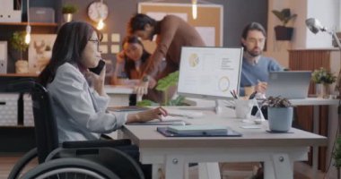 Tekerlekli sandalyedeki engelli iş kadını cep telefonuyla konuşuyor ve dahil çalışma alanında bilgisayarla çalışıyor. Akıllı telefon iletişimi ve kapsayıcı çalışma alanı kavramı.