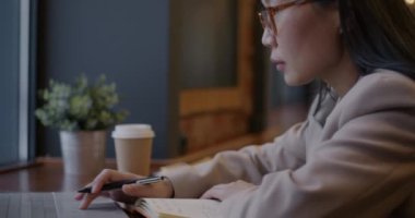 Kafede çalışan Asyalı iş kadını dizüstü bilgisayara bakıyor ve kahve içiyor. İş kadını öğle yemeği molasında işe odaklandı.