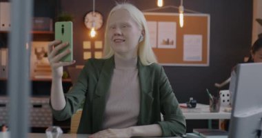 Neşeli albino iş kadını konuşuyor. Akşamları ofiste akıllı telefon ve kablosuz kulaklıkla Onlin 'i arıyor. İletişim ve çalışan kavramı.
