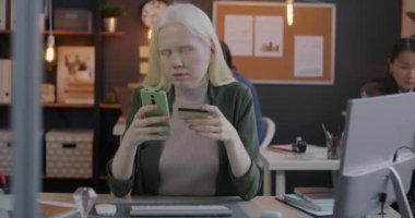 Akıllı telefon ve kredi kartı kullanan genç albino iş kadını ofiste para transferi yapıyor. Finans ve başarılı iş kavramı.