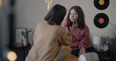 Anne, genç kızına makyaj malzemesi için yardım ediyor. Yatakta otururken ruj sürüyor. Annelik ve güzellik kavramı.
