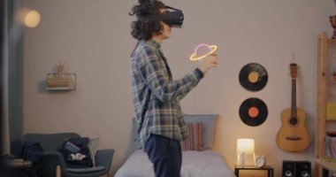 Sanal gerçeklik gözlüğü takan bir genç yatak odasında yürüyor ve modern aletlerle eğleniyor. Gelişmiş gerçeklik gözlükleri ve ergen çocuk konsepti.