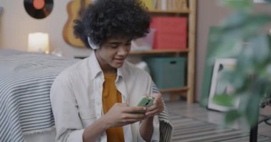 Neşeli Afrikalı Amerikalı ergen kulaklıkla müzik dinliyor ve yatak odasında akıllı telefon kullanıyor. Gençlik kültürü ve alet konsepti.