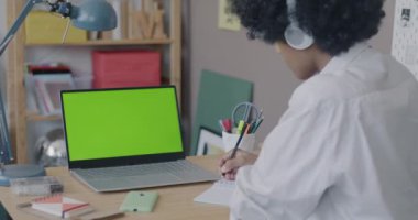 Çevrimiçi çalışan Afro-Amerikalı genç çocuk kroma tuşlu yeşil ekran laptopuna bakıyor kulaklıkla dinliyor ve evdeki masada yazıyor. Sanal eğitim ve evde eğitim kavramı.