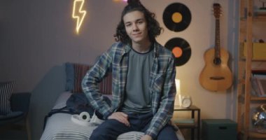 Akşamları yatakta oturmuş kameraya bakarak gülümseyen melez bir gencin ağır çekimde portresi. Gençlik ve ev kavramı.