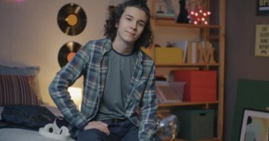 Kendine güvenen, şık kıyafetler giyen, akşamları yatak odasında kameraya bakan bir gencin yavaş çekim portresi. Ergenlik ve ev kavramı.