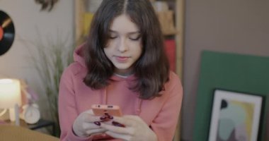 Akıllı telefon tutan neşeli kız ekranlara dokunuyor ve gülümsüyor evde yatakta oturuyor. Gadget ve gençlik yaşam tarzı konsepti.