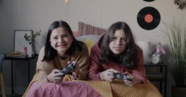 Yetişkin kadın ve genç kızın, yatak odasındaki kameraya bakarak konsol ve joystick ile heyecan verici video oyunları oynamalarının portresi. Aile ve eğlence konsepti.