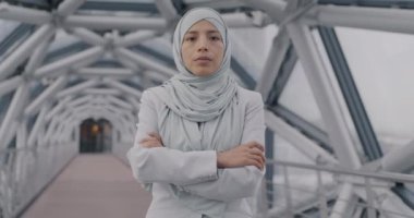 Geleneksel tesettürlü Orta Doğulu bir iş kadınının yavaşça portresi modern cam salonda duruyor ve kolları çaprazlanmış kameraya bakıyor. İş ve Müslüman kadın kavramı.