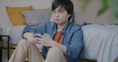 Akıllı telefon kullanan ve gülümseyen neşeli Asyalı adam yatak odasında oturmuş sosyal medyadan keyif alıyor. Gadget ve insanlar konsepti.