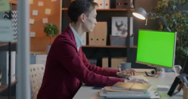 Çift ırklı genç iş adamı ofiste monitöre bakarak krom anahtar yeşil ekran bilgisayar kullanıyor. Modern teknoloji ve çalışan kavramı.