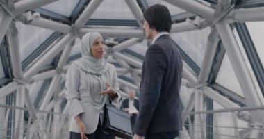 Asyalı iş adamı ve Orta Doğulu tesettürlü iş kadını çağdaş cam salonda el sıkışıyor ve konuşuyor. İş ve işbirliği kavramı.