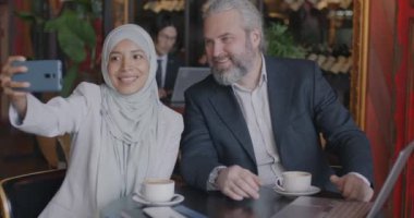 Orta Doğulu tesettürlü iş kadını Kafe 'deki masada akıllı telefon kamerası kullanarak Kafkas ortağıyla selfie çekiyor. İş ve fotoğraf konsepti.