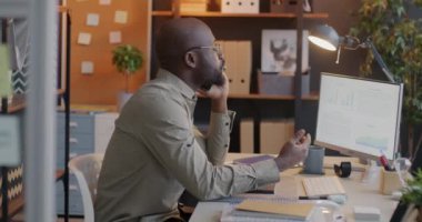 Cep telefonuyla konuşan Afro-Amerikalı işadamı ofiste çalışan bilgisayar ekranına bakıyor. Profesyonel iletişim ve iş kavramı.