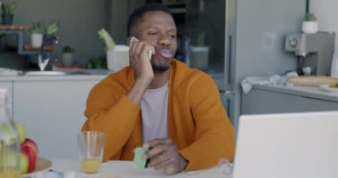 Cep telefonuyla konuşan mutlu Afro-Amerikalı adamın portresi mutfaktaki masada oturmuş gülümseyen haberleri tartışıyor. Cep telefonu ve iletişim kavramı.