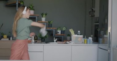 Modern bir evde, mutfakta kulaklıklarla dans ederek eğlenen neşeli genç bir bayan. Gençlik kültürü ve eğlence etkinliği konsepti.