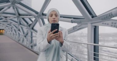 Modern cam duvar koridorunda duran akıllı telefonuyla çalışan kendinden emin Orta Doğulu iş kadınının yavaş hareketi. İş kadını ve mobil uygulama kavramı.