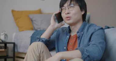 Neşeli Asyalı adam cep telefonuyla konuşuyor. İyi haberleri tartışıyor ve evde oturmuş gülümsüyor. İletişim ve insan kavramı.