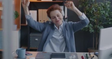 İş yerinde kulaklıkla müzik dinleyerek dans eden neşeli genç bayan ofis çalışanı. Modern kültür ve eğlence konsepti.