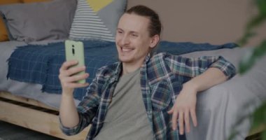 Neşeli adam akıllı telefonuyla online video görüşmesi yapıyor evde oturmuş el sallıyor. İletişim ve sanal buluşma kavramı.
