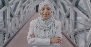 Modern cam salonda kameraya gülümseyen tesettürlü Orta Doğulu genç iş kadınının yavaş çekimde portresi. İş ve insan kavramı.