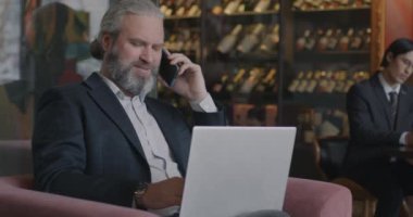 Laptopla çalışan deneyimli bir iş adamı ve kafedeki koltukta oturan cep telefonuyla konuşan biri. İş ve profesyonel iletişim kavramı.